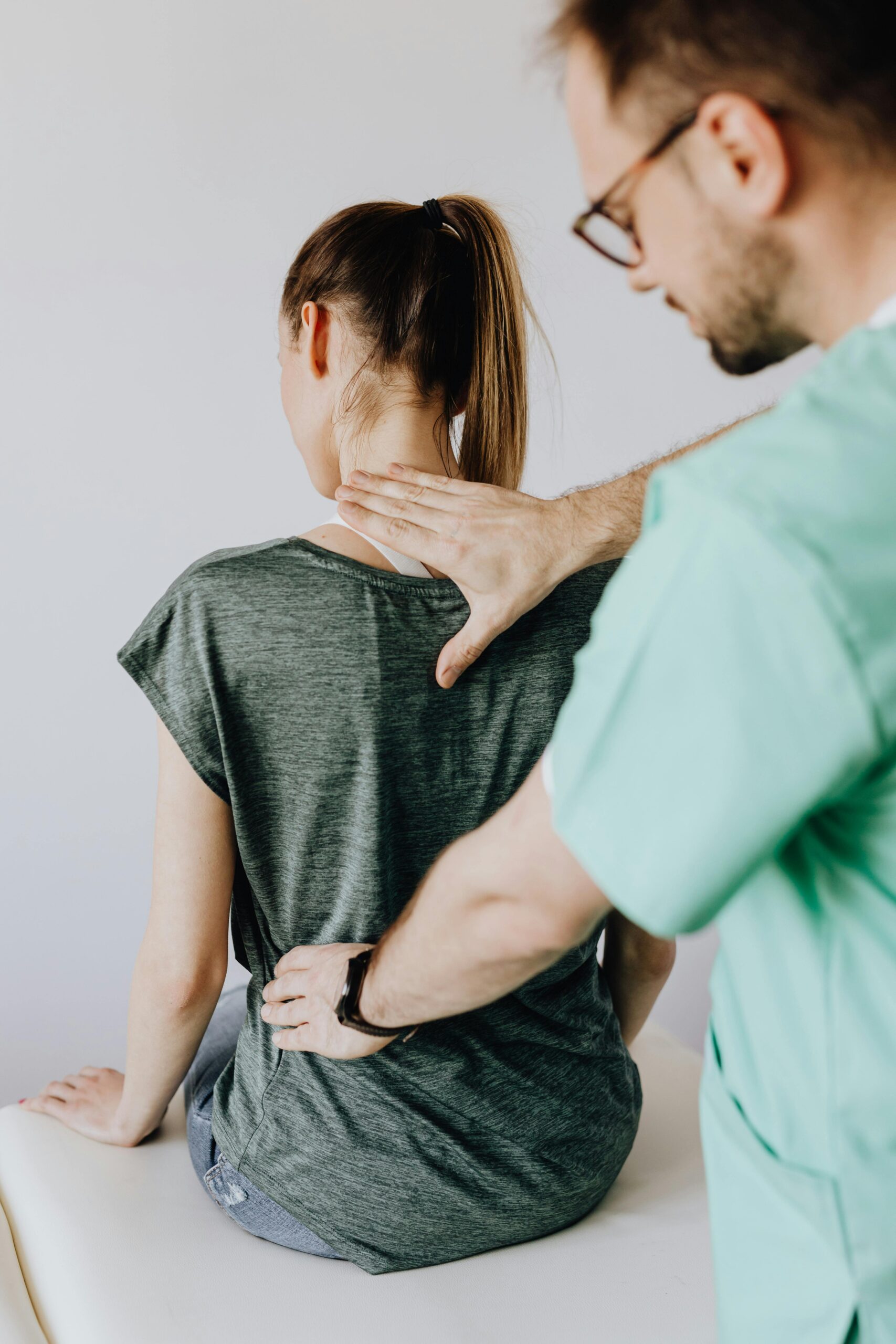 From above back view of crop orthopedist in uniform and eyewear examining back of slim faceless female in casual clothes sitting on examination couch in doctor office